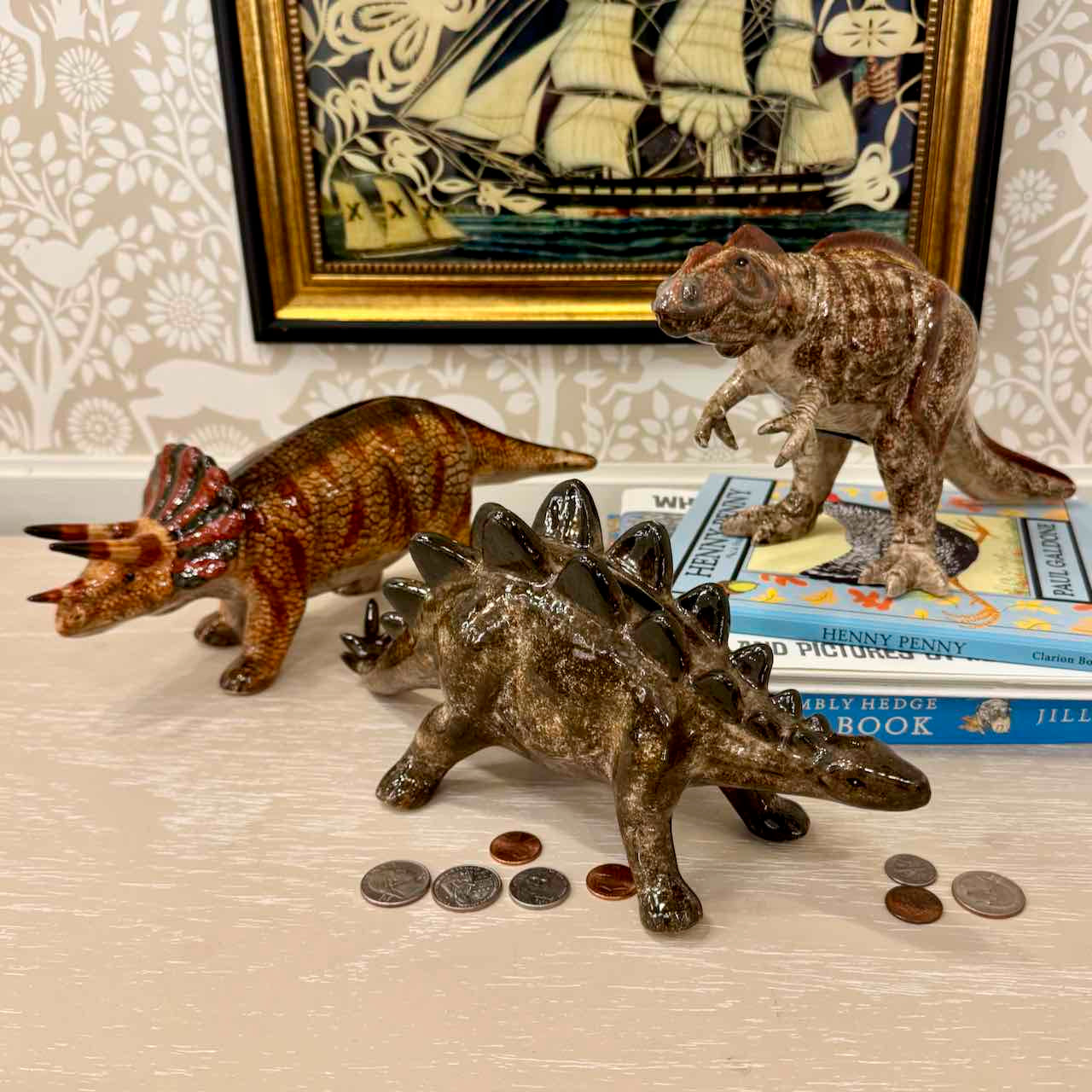Three dinosaur figurines on a surface with coins and books underneath, framed artwork in the background.