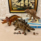 Three dinosaur figurines on a surface with coins and books underneath, framed artwork in the background.