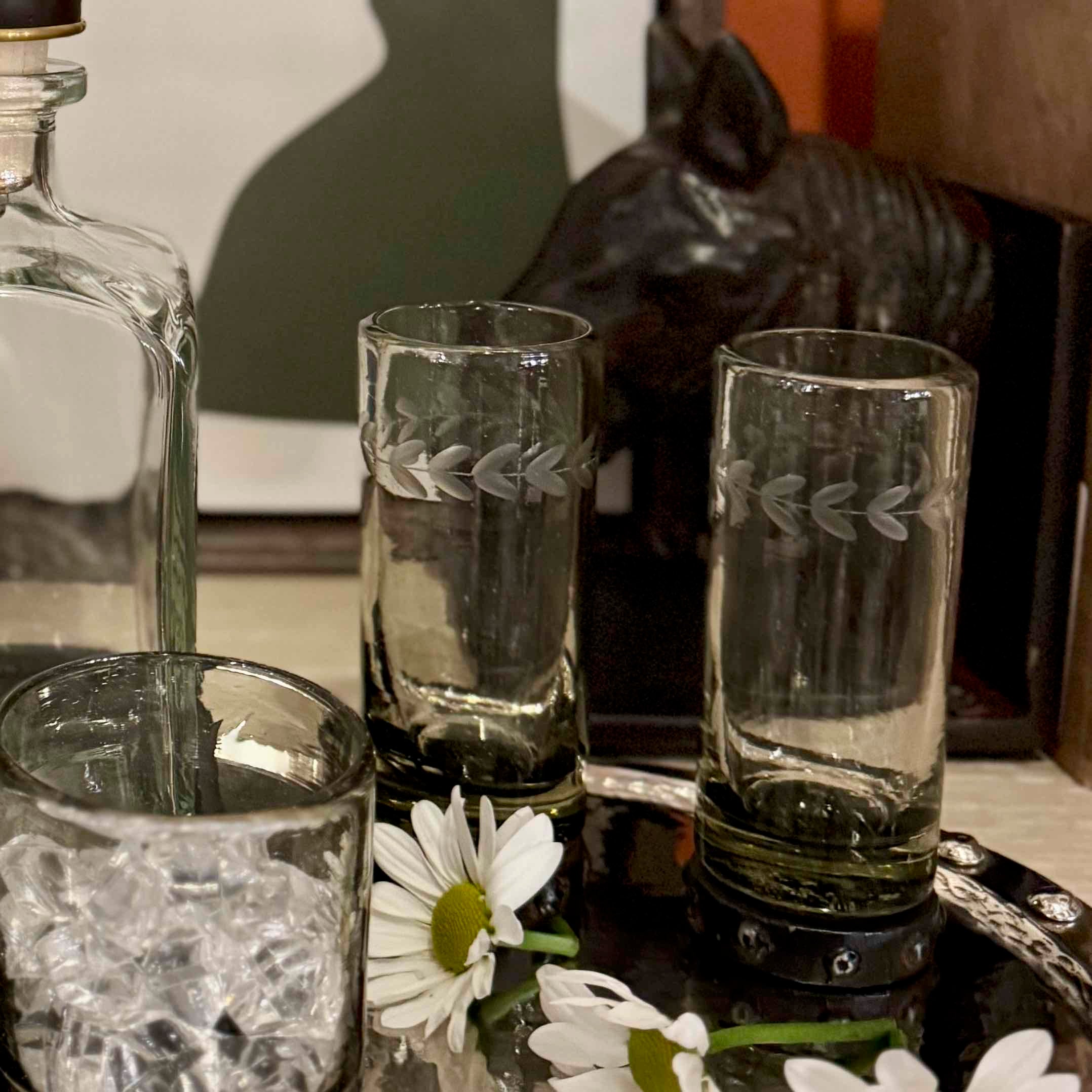 Set of etched glass tumblers on a reflective surface with flowers and a bottle.