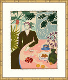 A framed art print featuring an illustration of a woman seated at a table with food and plants, surrounded by a wide white mat and ornate gold frame.
