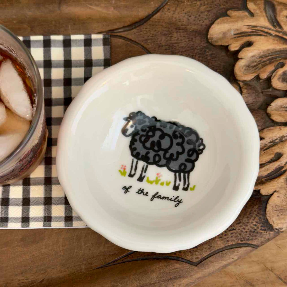 A white ceramic bowl with a scalloped rim, featuring a black cartoon sheep illustration and the text 'of the family' in black script. The bowl is styled on a rustic wooden surface next to a checkered napkin and a glass of iced tea.