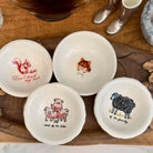 Four small white ceramic bowls with animal illustrations and text, arranged on a wooden tray. The bowls feature drawings of a red squirrel, a brown cat, a pink pig family, and a black sheep, with phrases like 'Don't touch my nuts' and 'most of the litter' and 'most of the family'.