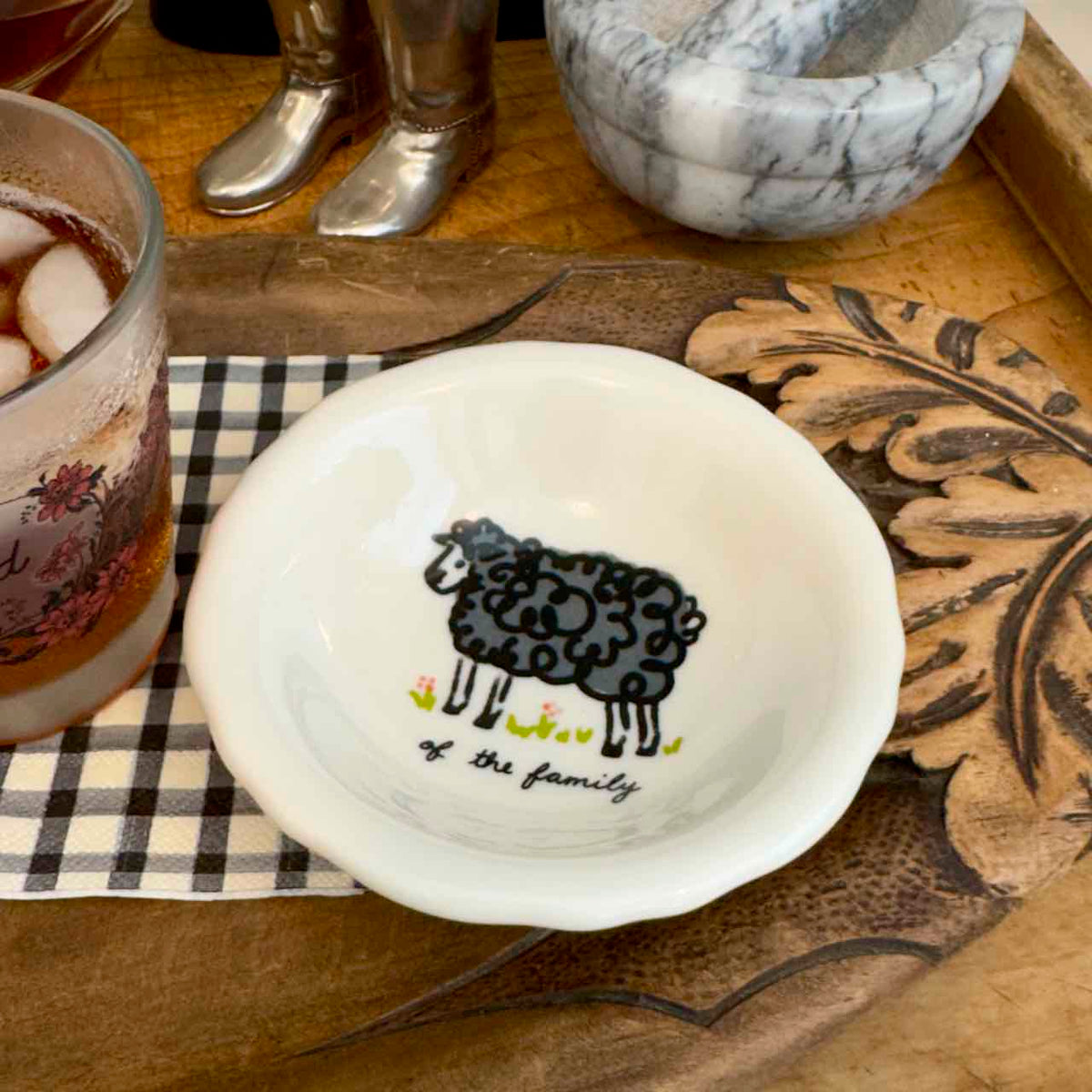 A small white ceramic bowl with a black sheep illustration and the text 'of the family' inside, styled on a wooden tray with a checkered napkin, a drink, and other tabletop objects.