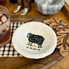 A small white ceramic bowl with a black sheep illustration and the text 'of the family' inside, styled on a wooden tray with a checkered napkin, a drink, and other tabletop objects.