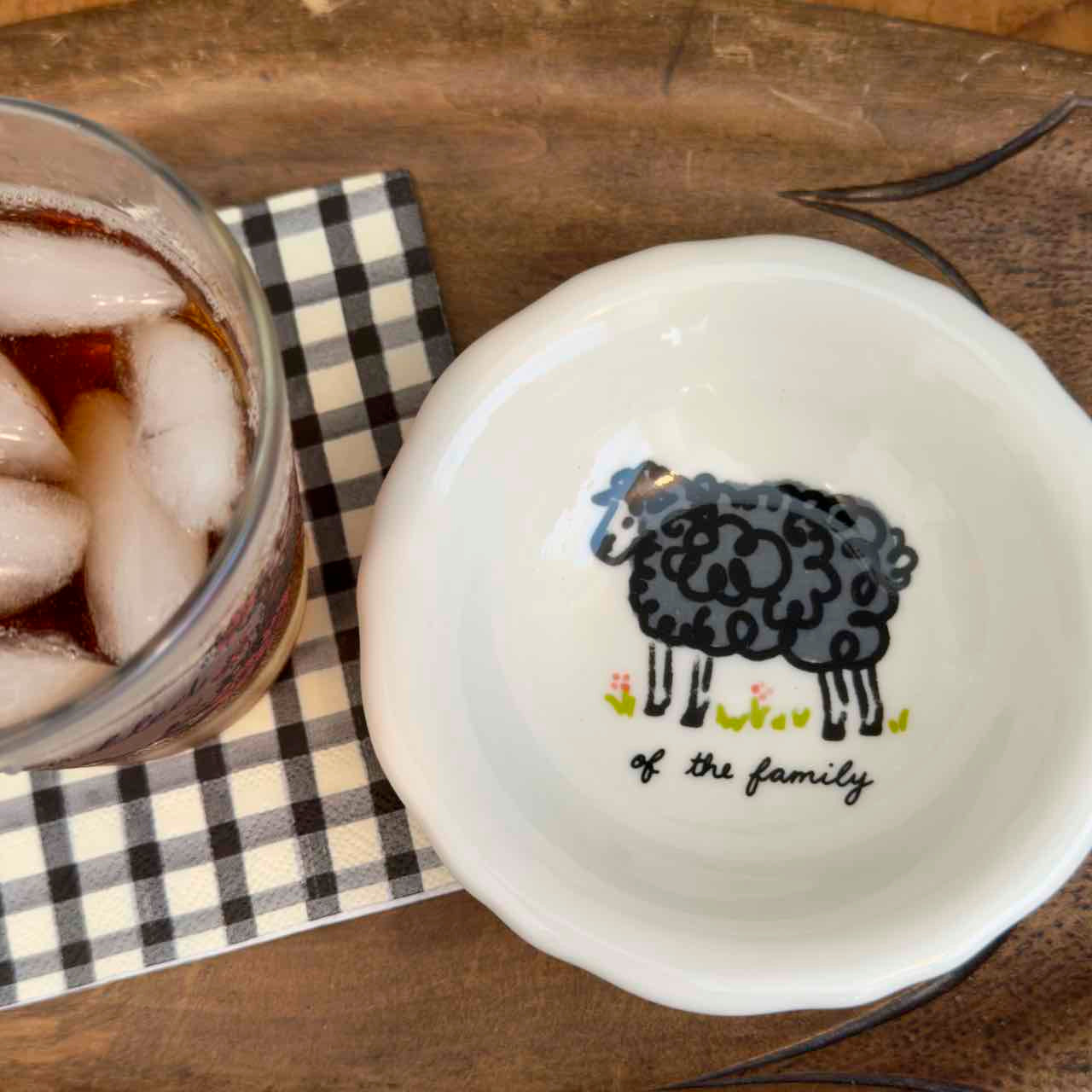 A white ceramic bowl with a black sheep illustration and the text 'of the family' printed inside, shown on a wooden surface next to a glass of iced soda on a black-and-white checkered napkin.