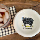 A white ceramic bowl with a black sheep illustration and the text 'of the family' printed inside, shown on a wooden surface next to a glass of iced soda on a black-and-white checkered napkin.