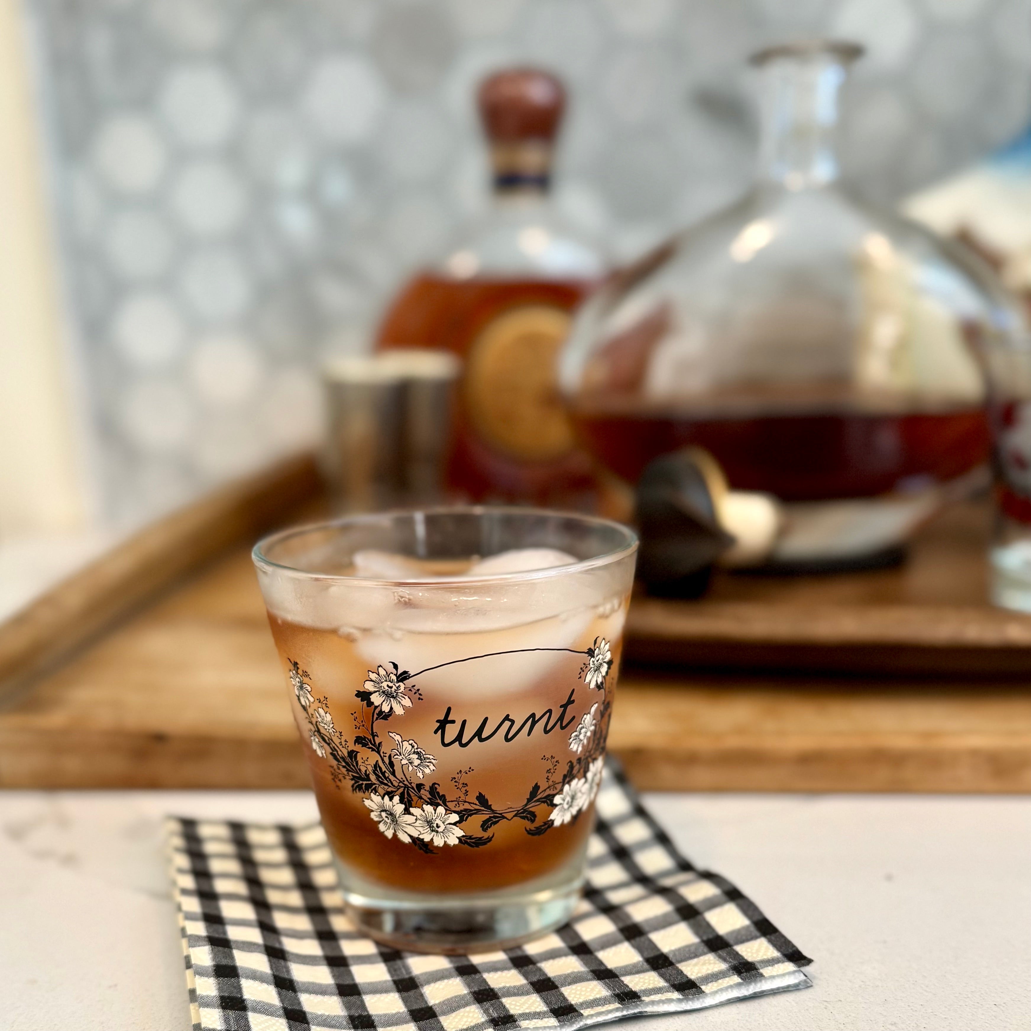 A clear rocks glass filled with an amber drink and ice, resting on a black-and-white checkered napkin. The glass has a floral design and the word 'turnt' printed on it.