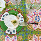 A top-down view of a cheese wedge on a green and white ceramic plate with blue floral accents, set on a colorful patterned tablecloth with wine glasses nearby.