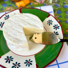 A round wheel of soft white cheese, with one slice removed, resting on a decorative green and white plate with blue flowers. A small metal cheese server with a black handle is placed on the slice.