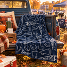 A navy blue fleece throw blanket with white football-themed doodles and 'GO TEAM' pennant graphics is draped over the back of a rustic blue pickup truck. The scene is set outdoors at an autumn market with fall foliage, pumpkins, a thermos, and camping chairs in the background.