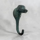 A decorative hanging hook shaped like a dog's head, finished with a dark green patina and blackened highlights, set against a light gray textured background.