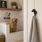 A golden retriever sits in a white tiled pet bathtub while a gray towel hangs from a dark green dog-head wall hook in an adjacent bathroom doorway.
