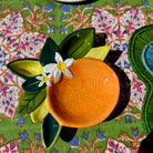 Ceramic bowl shaped like an orange with leaves and a flower on a patterned tablecloth.