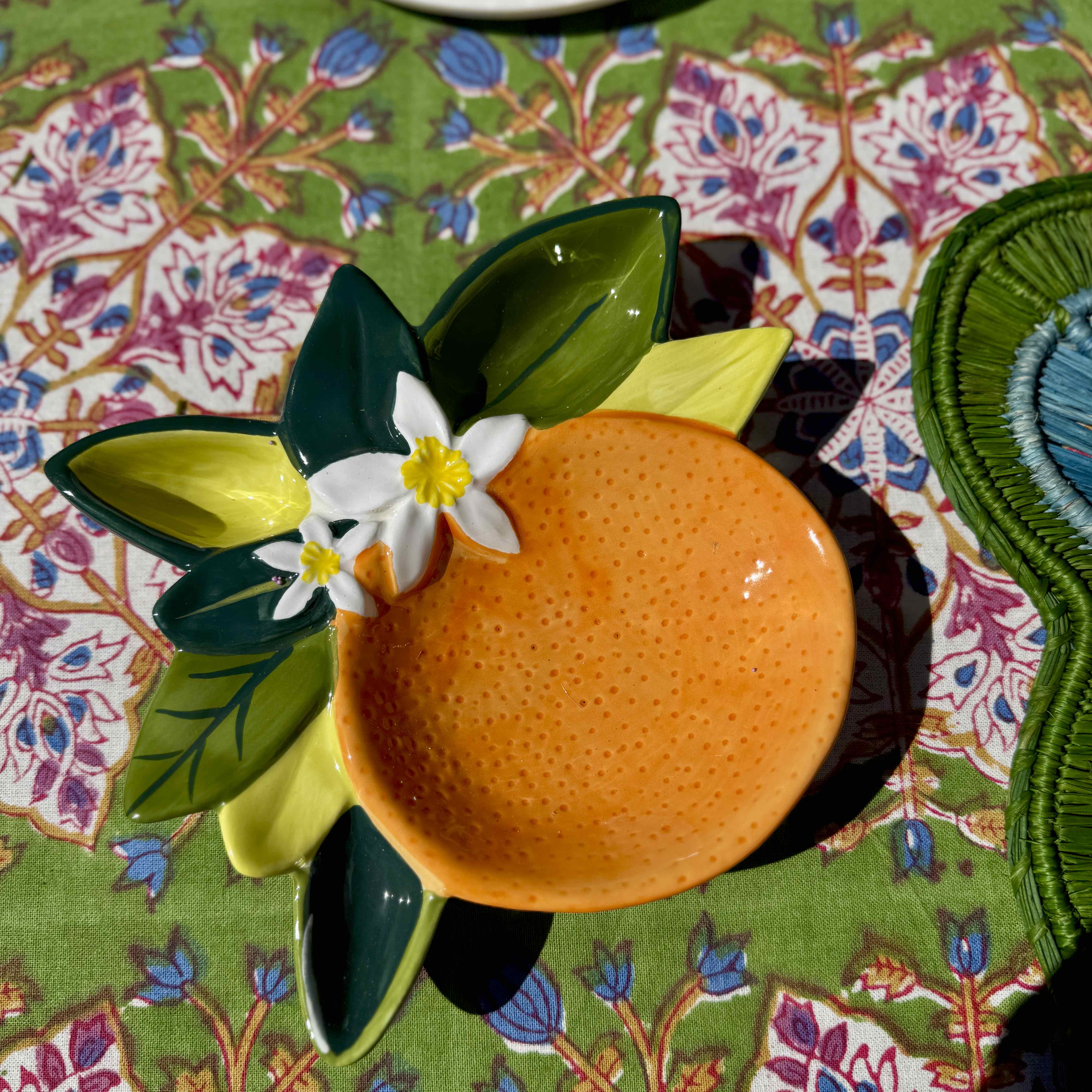 Ceramic bowl shaped like an orange with leaves and a flower on a patterned tablecloth.