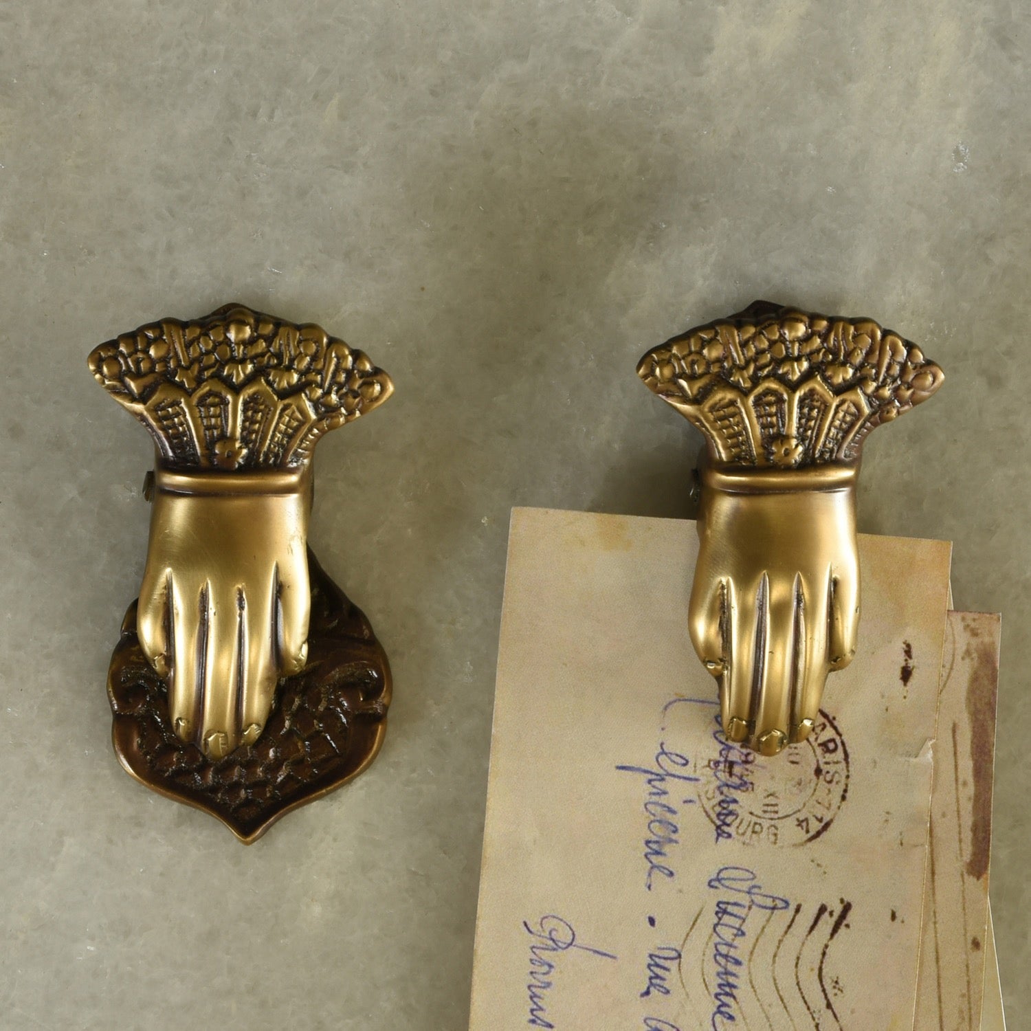 Two antique-style brass door pull handles shaped like hands holding a bouquet of flowers, mounted on a textured gray wall next to a vintage handwritten postcard.