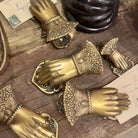A set of ornate antique-style brass hand-shaped drawer pulls with intricate filigree cuffs, arranged on a rustic wooden surface alongside vintage papers.
