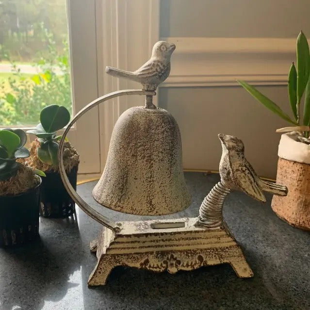 Decorative bell with bird sculptures on a surface near potted plants