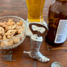 Cowboy boot bottle opener with a glass of beer, bowl of nuts, and bottle in the background on a wooden surface.
