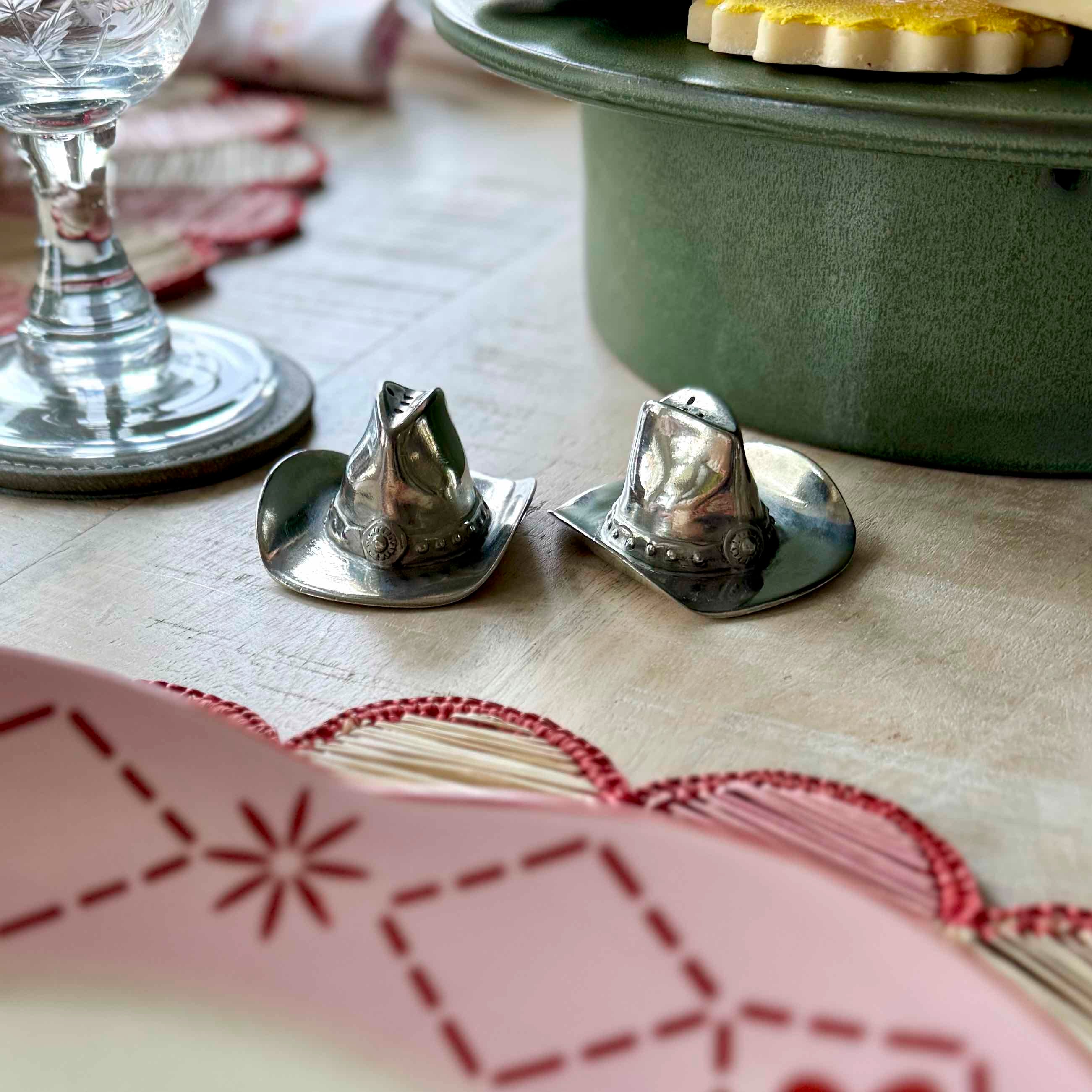 Two silver salt shakers on a table with a green pot and glass in the background.