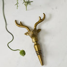 A brass deer head bottle stopper with antlers and a pointed pourer base, styled on a white marble surface with greenery and a bottle in the background.
