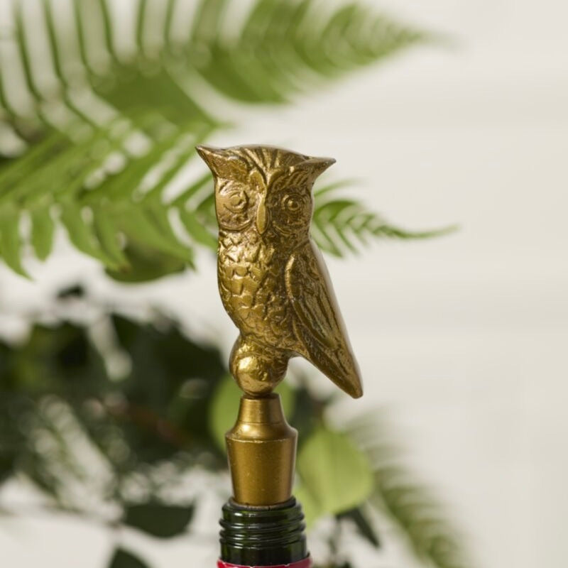 A close-up of a gold-toned metal owl-shaped bottle stopper resting on the neck of a dark bottle, with blurred green fern leaves in the background.