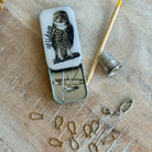 Small metal tin with owl design on a wooden surface with sewing tools.