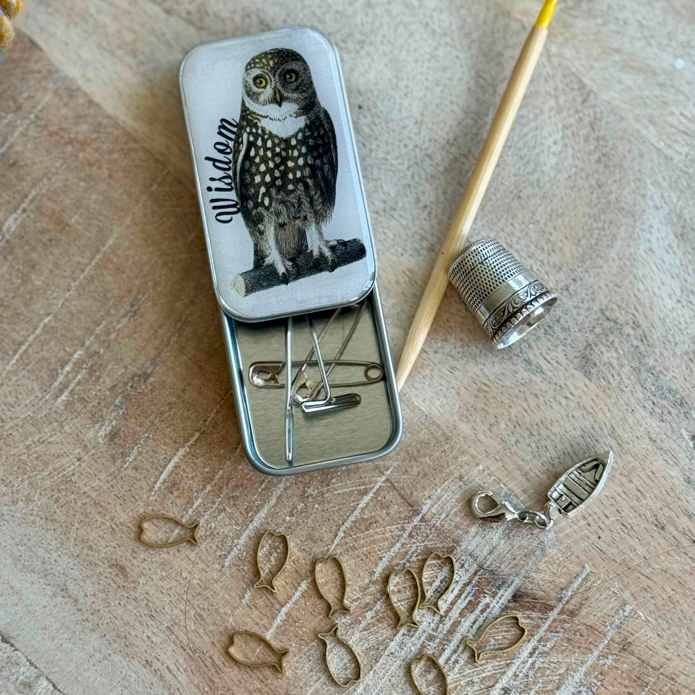 Small metal tin with owl design on a wooden surface with sewing tools.