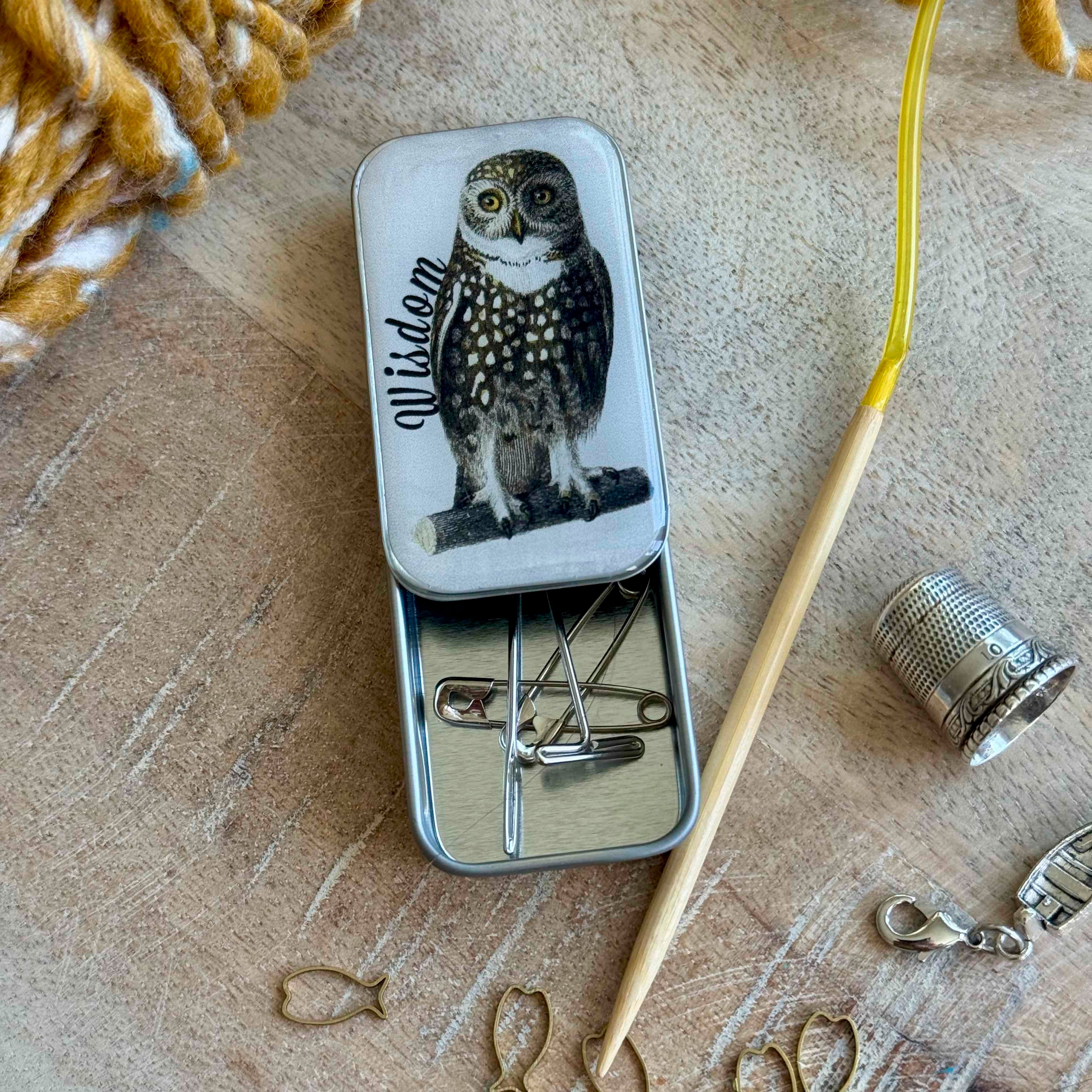 Small metal tin with owl design on lid, containing sewing needles, on a wooden surface with yarn and thimble.