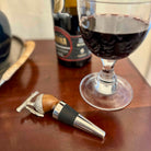 Wine bottle stopper with wooden handle and metal tip next to a glass of red wine on a wooden surface.