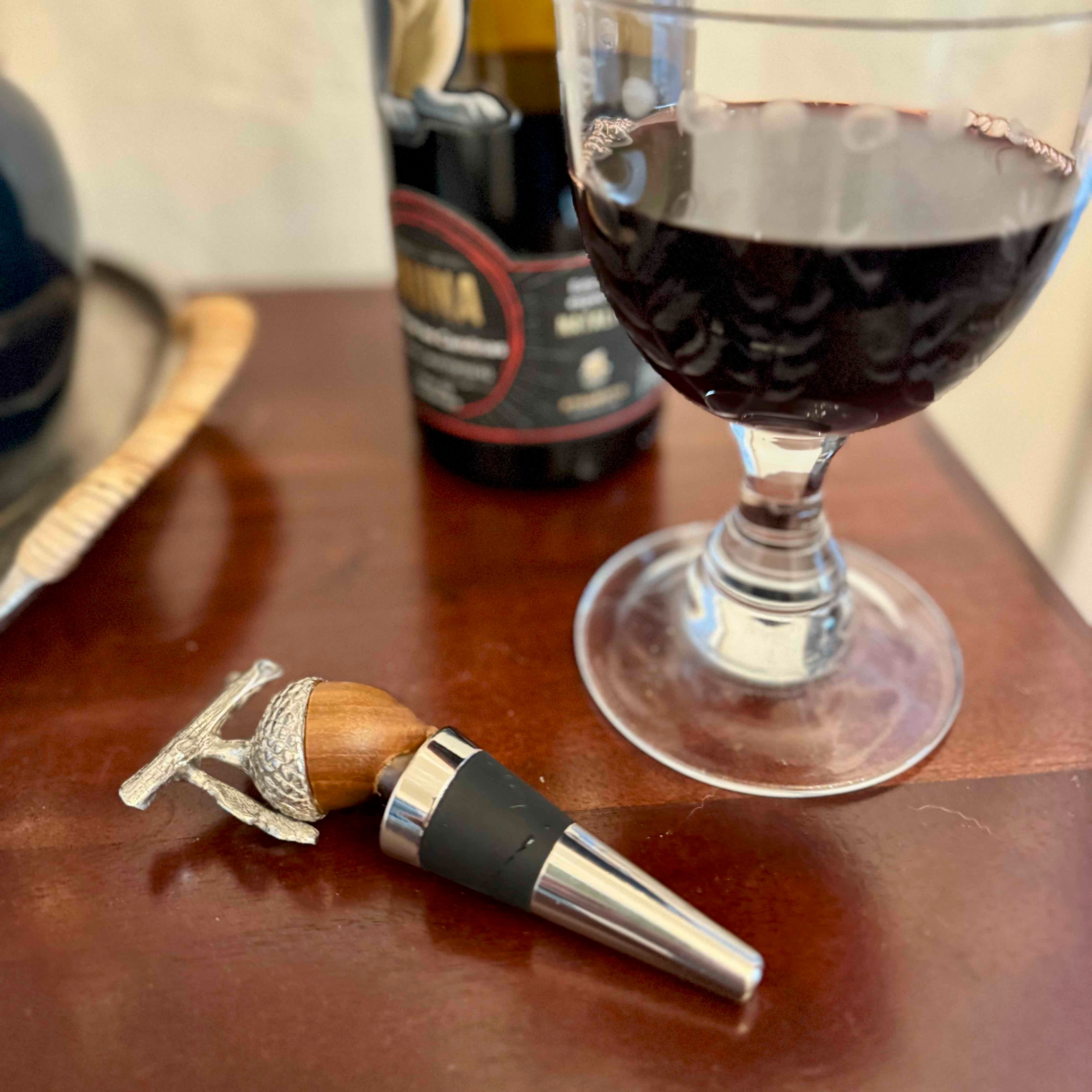 Wine bottle stopper with wooden handle and metal tip next to a glass of red wine on a wooden surface.
