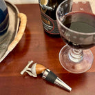 Wine bottle stopper with wooden handle and metal accents on a wooden surface with a glass of red wine and a bottle in the background.