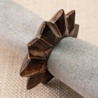 A carved dark brown wooden napkin holder with a star-like geometric design, shown holding a rolled light gray linen napkin on a textured beige surface.