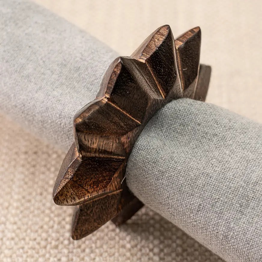 A carved dark brown wooden napkin holder with a star-like geometric design, shown holding a rolled light gray linen napkin on a textured beige surface.