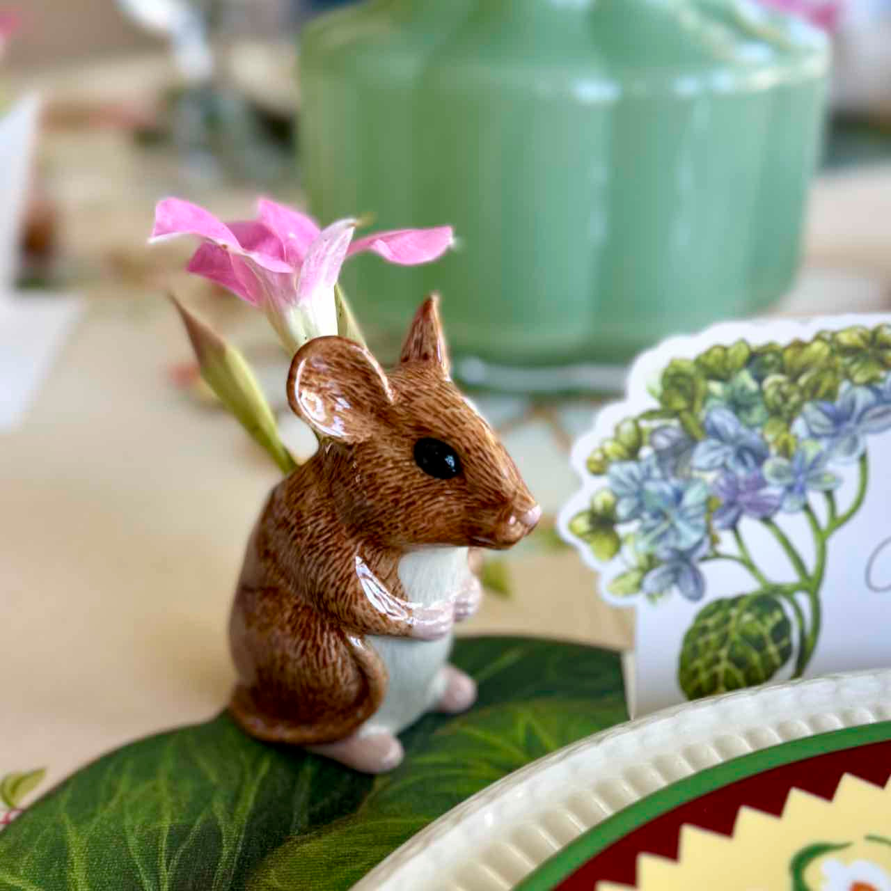 Small figurine of a mouse on a decorative plate with a floral teacup in the background.