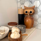 Wooden owl figurine on a shelf with decorative items against a tiled wall.