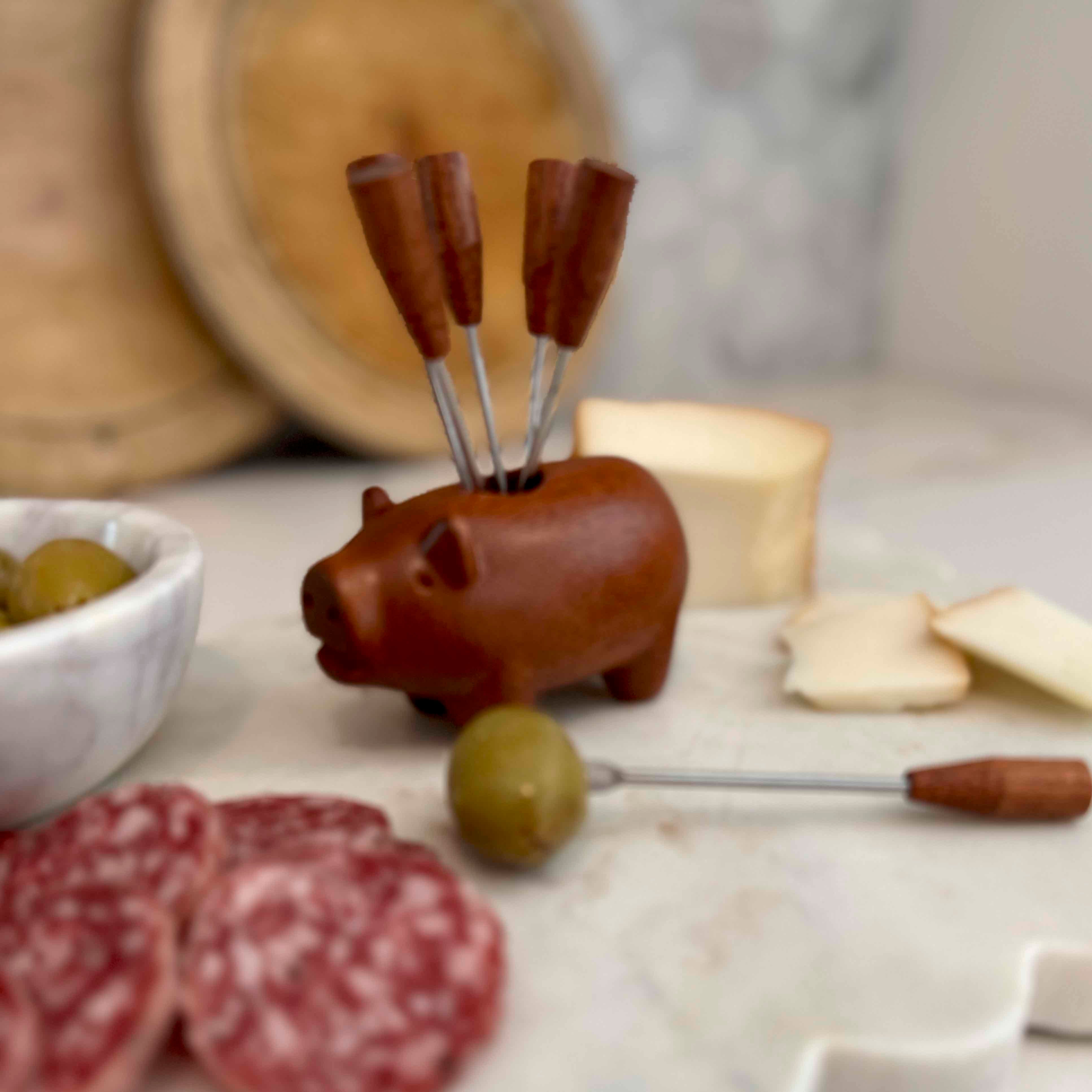 Pig-shaped appetizer set with skewers on a table with olives, salami, and bread.