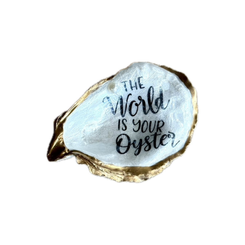 A novelty oyster shell pin or brooch featuring the hand-lettered quote 'THE World IS YOUR Oyster' in black ink on a textured white interior, framed by a metallic gold-rimmed edge, shown on a plain white background.