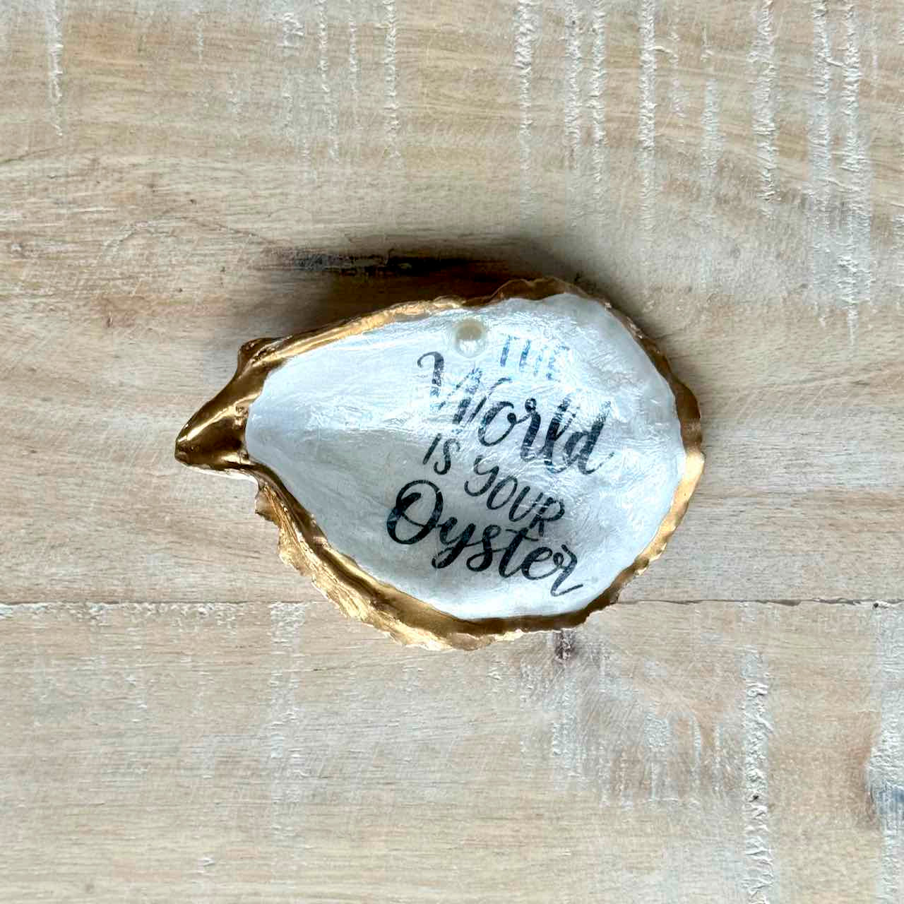 A hand-painted oyster shell decorative piece with gold-painted edges and a white interior featuring the hand-lettered quote 'The World Is Your Oyster', displayed on a light wood background.