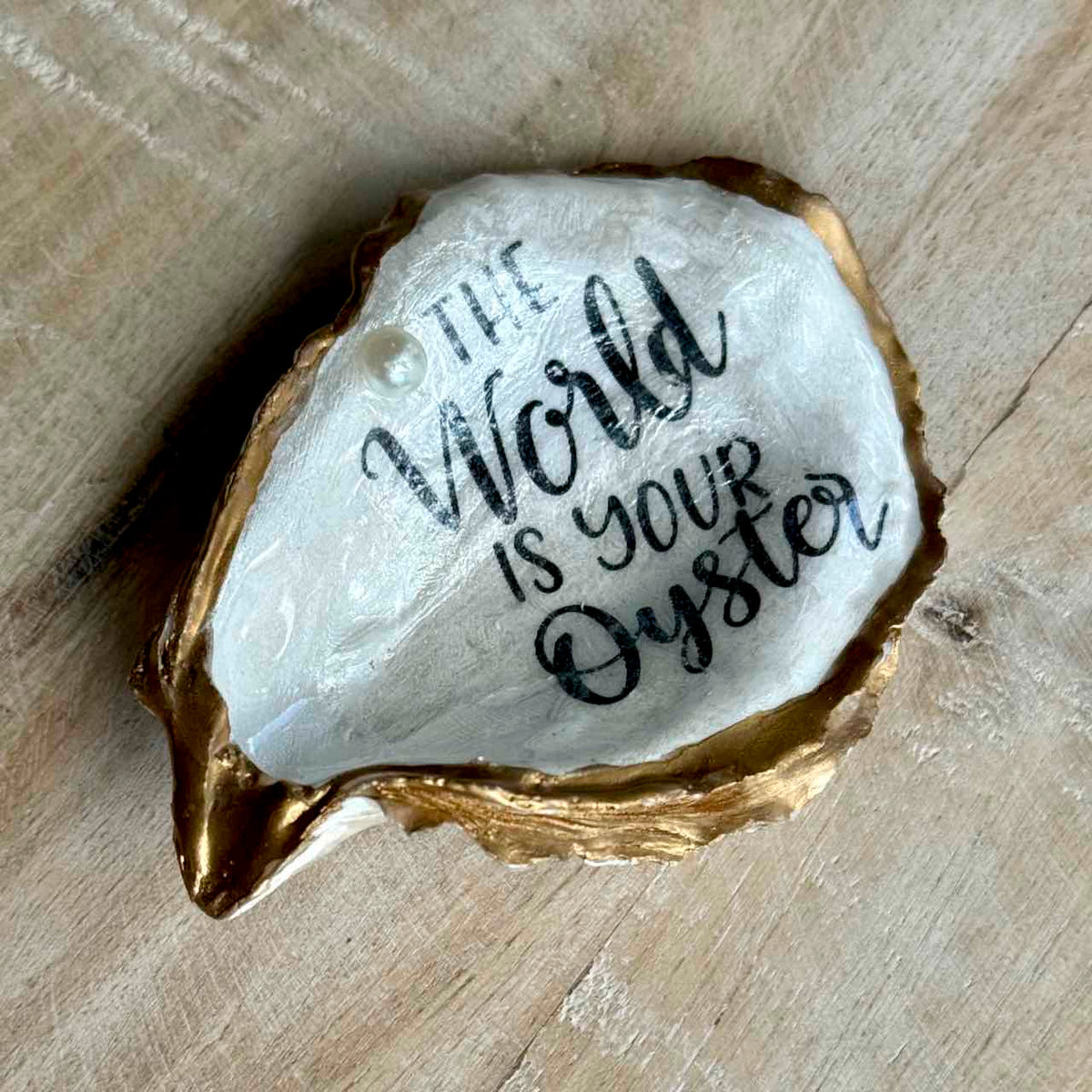 A hand-painted oyster shell art piece with a gold-toned rim and a white interior, featuring the hand-lettered quote 'THE World IS YOUR Oyster' in black ink, displayed on a light wood background.