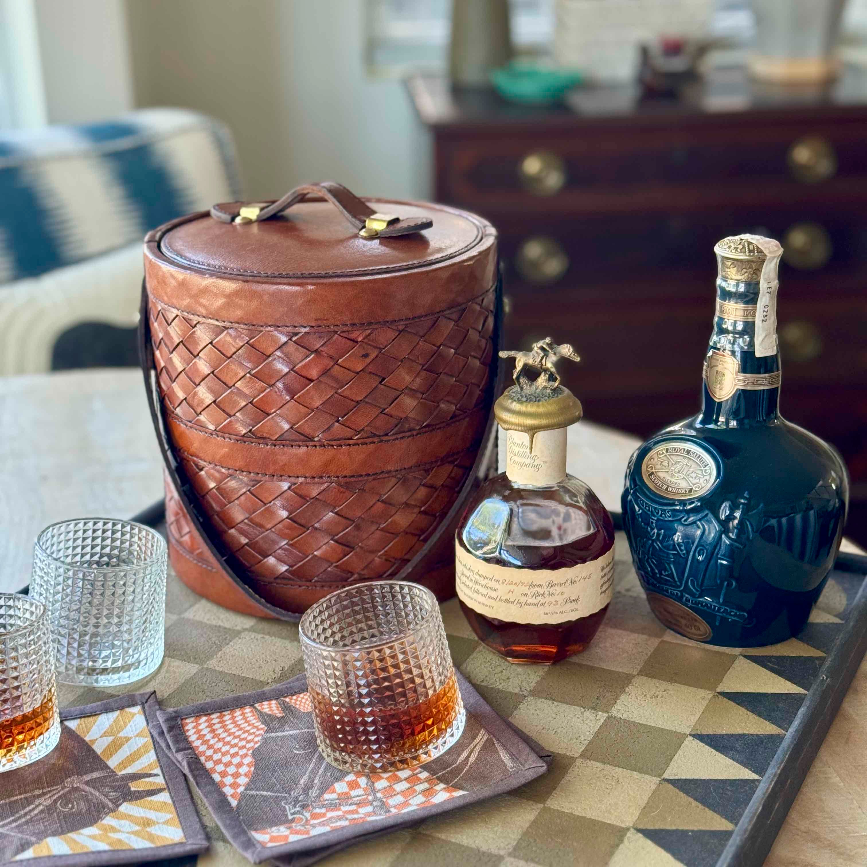 Beverage setup with bottles, glasses, and a woven leather cooler on a checkered tablecloth.