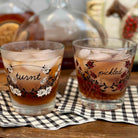 Two decorated rocks glasses filled with an amber drink and ice, placed on a checkered napkin on a wooden table with blurred bottles in the background. One glass is labeled 'turnt' with white flowers and the other 'pickled' with red flowers.
