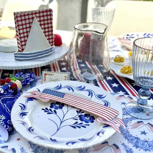 Patriotic-themed table setting with a blue floral plate, red and white striped napkin, American flag place card, and matching red, white, and blue table linens.