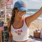 Woman wearing a 'USA' tank top and blue cap with an American flag, standing on a beach with July 4th decorations.